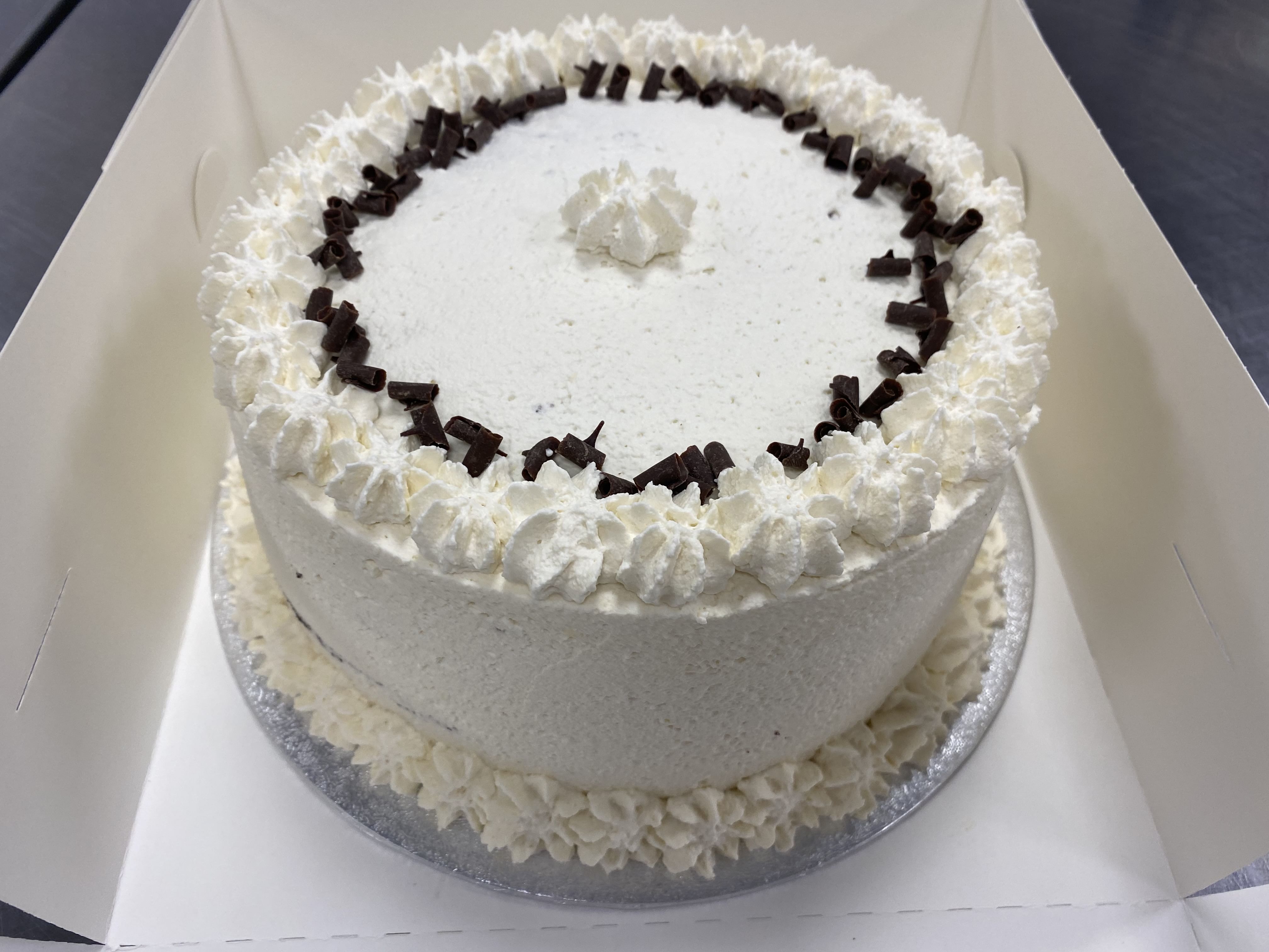 A black forest gateaux, round and delicately frosted white, arrives in a box. It features intricately piped white icing around the edge and a central rosette. Dark chocolate shavings elegantly encircle the top, just inside the white icing border. The cake is presented on a silver board and enclosed within pristine white cardboard.